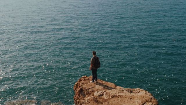 Man standing on rocky cliff facing vast ocean with backpack and sunlit water glinting