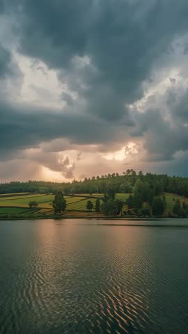 Vertical sunbeam video piercing storm clouds, illuminating farmland and reflective lake