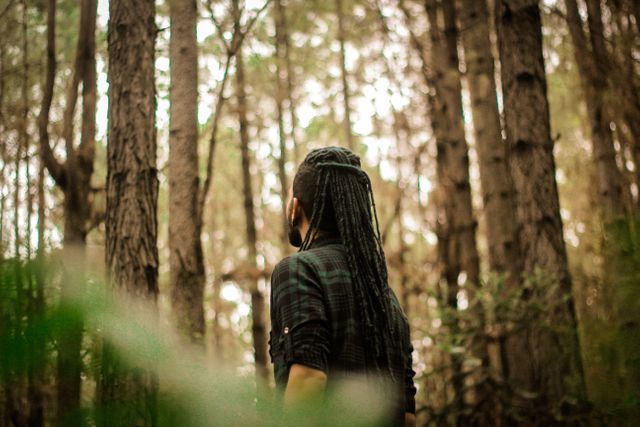 Man with dreadlocks seen from the back, surrounded by tall trees in dense forest, suggesting themes of adventure, nature exploration, and solitude. Useful for articles, blogs, and advertisements focusing on outdoor activities, nature, tranquility, personal journeys, or eco-friendly lifestyles.