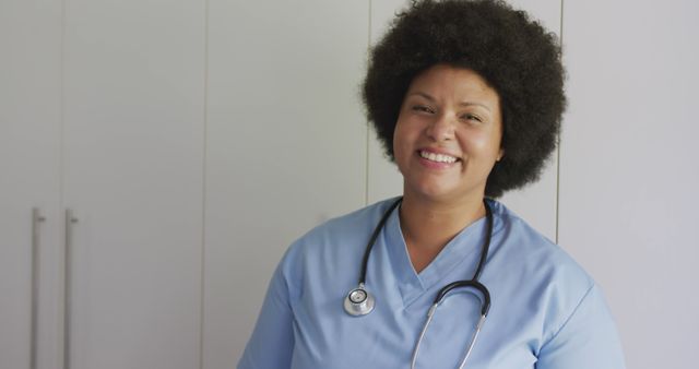 African American nurse wearing blue scrubs and stethoscope, smiling in clinical setting. Ideal for depicting healthcare professionals' dedication, medical services, patient care, hospital staff, and friendly medical assistance visuals. Suitable for healthcare advertisements, medical websites, and promotional health campaigns.