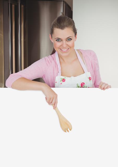 Woman cook stands in a modern kitchen, wearing a floral apron and holding a wooden fork while standing behind a blank poster. The blank space provides an excellent opportunity for adding custom text or graphics for advertisements, culinary promotions, recipes or food blogs.