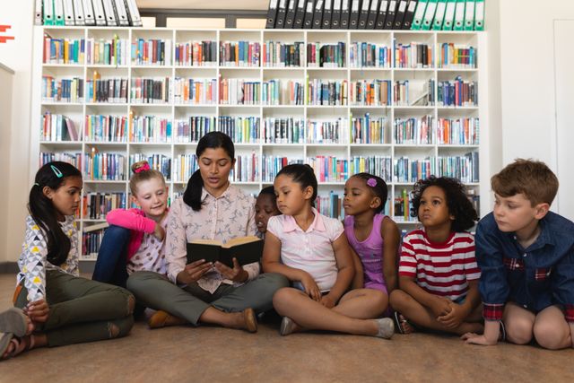 Female teacher reading a book to a diverse group of young students sitting on the floor in a school library. Shelves filled with books in the background. Ideal for educational content, school promotions, literacy programs, and multicultural learning materials.