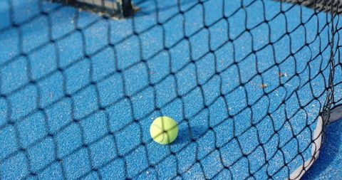 Tennis ball resting against net on vibrant blue court casting diamond mesh shadows