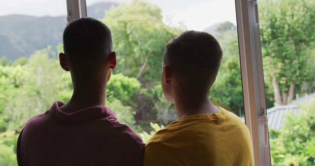 Rear view of biracial gay male couple standing looking out of window leaning heads together. staying at home in isolation during quarantine lockdown.