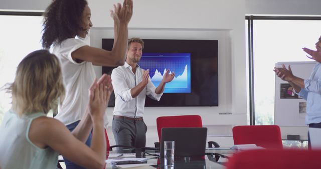 Diverse team members are seen standing and applauding during a business meeting. Background includes a large screen displaying a positive trend graph. Suitable for concepts related to business success, teamwork, corporate milestones, professional collaboration, and office culture.