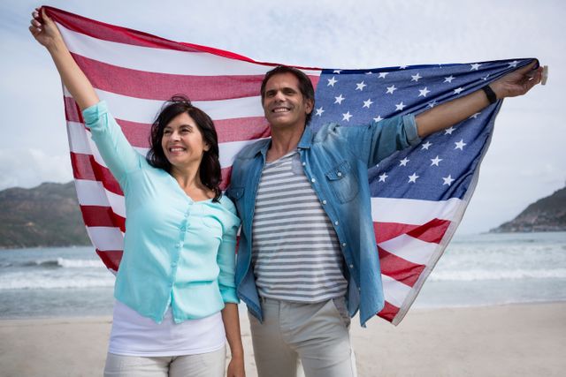 Couple standing on beach holding American flag, smiling and enjoying winter day. Ideal for themes of patriotism, holidays, travel, and outdoor activities. Perfect for use in advertisements, travel brochures, and social media posts celebrating national pride and togetherness.