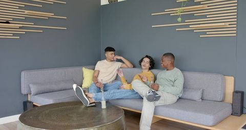 Multicultural young men chatting and relaxing on modern gray sofa with drinks