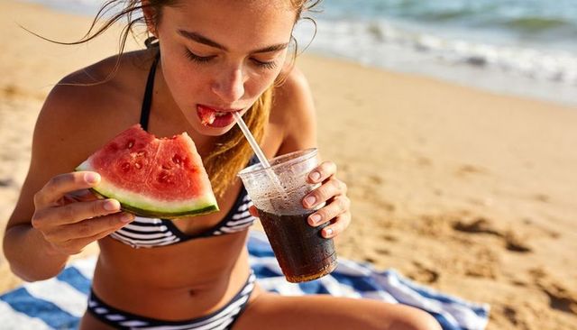 Young woman enjoying watermelon and sipping cola on sunny sandy beach in striped bikini