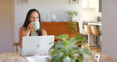 African American woman sipping coffee while working on laptop in sunlit minimalist home