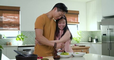 Asian couple embracing while preparing fresh avocado salad at modern home kitchen island