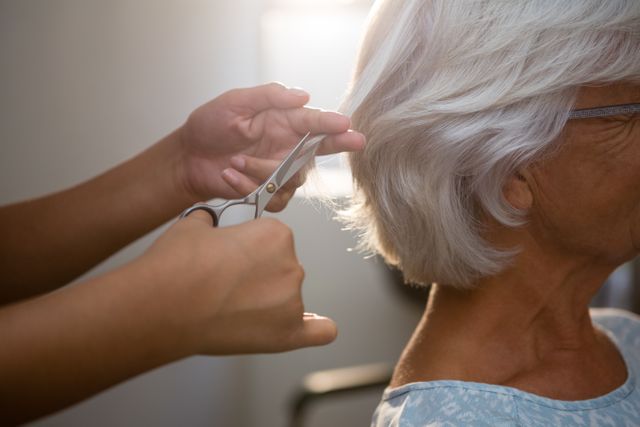 Beautician cutting senior woman's hair in a salon, focusing on the hands and scissors. Ideal for use in articles or advertisements related to hair care, beauty services, senior grooming, and professional hairstyling.