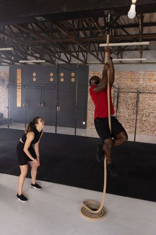 Male athlete climbing rope while female coach spotting in industrial brick gym training