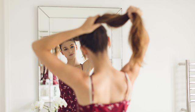 Woman tying her hair in front of a bathroom mirror, reflecting her image. She is wearing a red dress with floral patterns. Ideal for use in articles or advertisements related to beauty, personal care, morning routines, and lifestyle content.