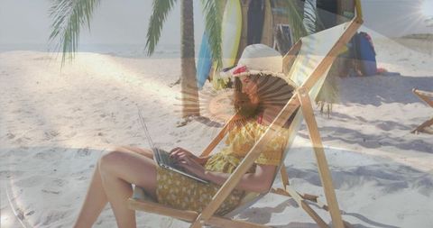 Working remotely on tropical beach, woman reclining in yellow dress using laptop