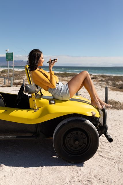This image depicts a smiling woman sitting on a yellow beach buggy, using her smartphone by the sea on a sunny day. Ideal for travel and adventure promotions, summer holiday advertisements, and lifestyle blogs focusing on leisure and relaxation.