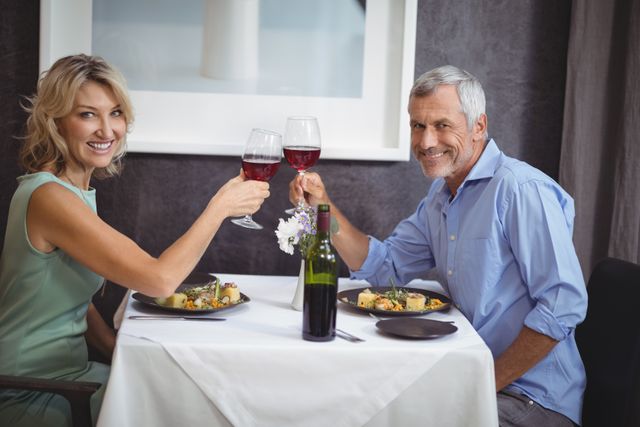 Mature couple toasting glasses of red wine while enjoying a meal in an elegant restaurant. Perfect for themes of romance, celebrations, anniversaries, or marketing fine dining experiences.