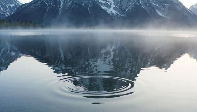 Rippling concentric rings on glassy alpine lake reflecting snow-capped peaks at sunrise
