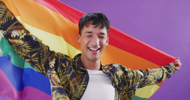 Young man holding a rainbow flag with a happy smile on his face against a purple background. Ideal for use in campaigns promoting LGBTQ+ rights, pride events, diversity and inclusion initiatives, and equality awareness. The vibrant colors and joyful expression convey a strong sense of celebration and support.