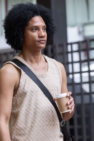 Urban man holding coffee cup and wearing beige tank with crossbody strap, standing and looking left
