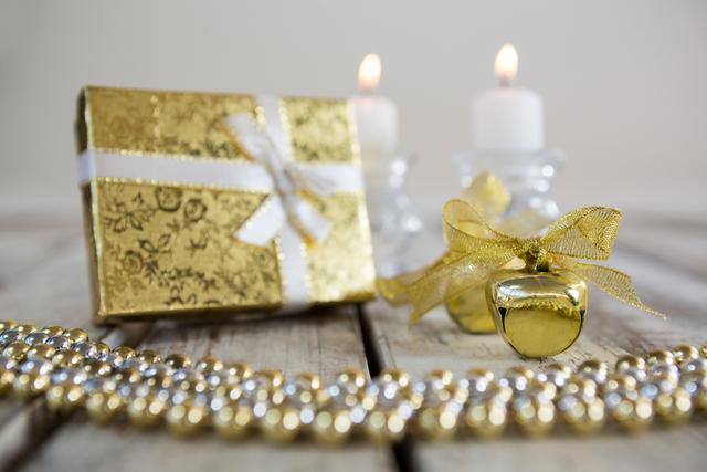 Christmas decorations including a gold gift box with a white ribbon, two lit candles, and gold ornaments on a wooden plank. Ideal for holiday greeting cards, festive advertisements, and seasonal blog posts. The warm and elegant setup evokes a traditional Christmas atmosphere, perfect for promoting holiday sales or decorating ideas.