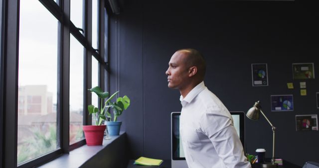 Business professional pondering while standing by window in modern office. This could be used in corporate websites, business strategy blogs, and inspirational business materials promoting productivity and leadership.