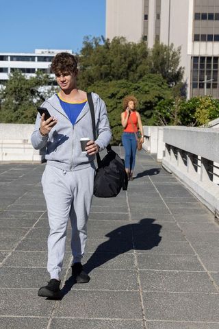 Young man walking checking smartphone holding coffee while woman walking behind on rooftop