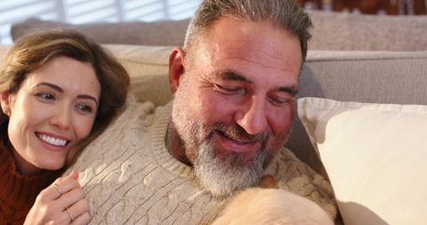 Mature couple cuddling on sofa in cable-knit sweaters petting small dog, cozy home moment