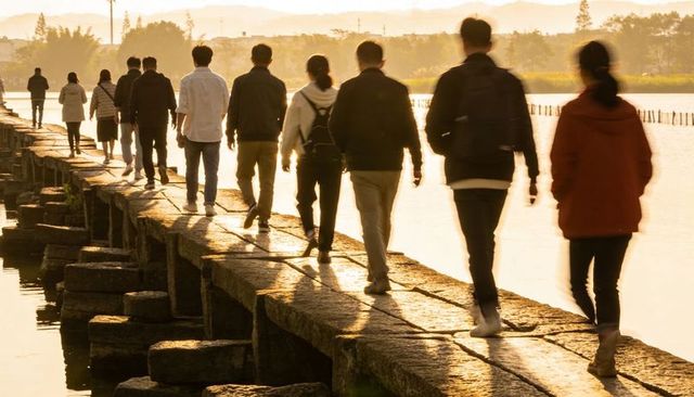 Walking group crossing golden-lit stone causeway over lake at sunset with backlit silhouettes