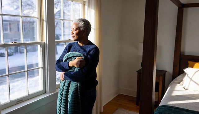 Senior woman gazing out snowy window holding teal blanket, peaceful sunlit bedroom moment