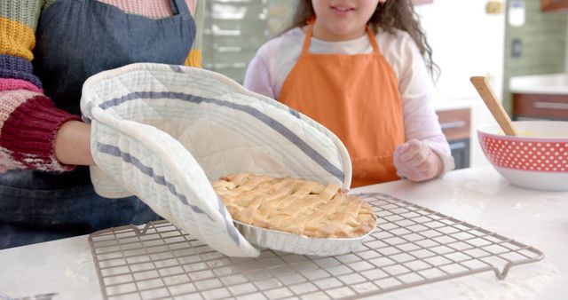 A mother and daughter use oven mitts to take a freshly baked apple pie out of the oven, placing it on a wire rack to cool. They smile and bond while enjoying their cooking activity in the kitchen. This image can be used for content relating to family bonding, home cooking, culinary activities, and promoting baking products or recipes.