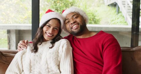 Smiling diverse couple wearing santa hats and cozy cable knit sweaters, sitting on couch