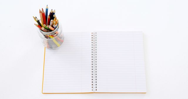 A glass jar filled with colorful pencils is placed next to an open blank notebook, with copy space. Ideal for educational themes, the setup invites creativity and note-taking.