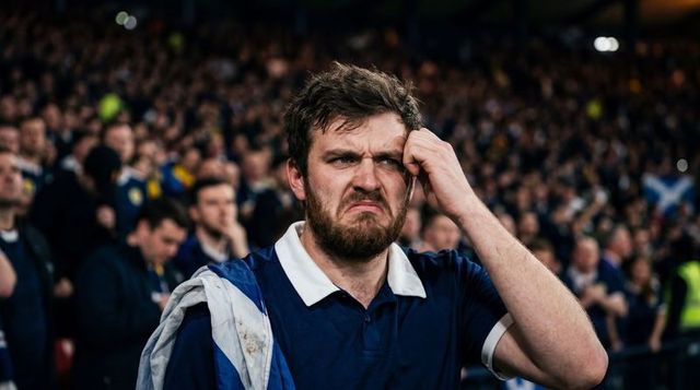 Grimacing football fan rubbing temple, wearing team flag and navy polo in stadium crowd