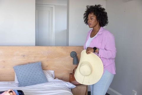 African american woman packing for trip holding straw sun hat in minimalist hotel room