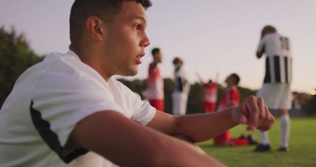 A young male soccer player in sportswear is resting after an intense match at sunset, with teammates in the background. Useful for depicting themes of sports, teamwork, determination, and relaxation after physical exertion.