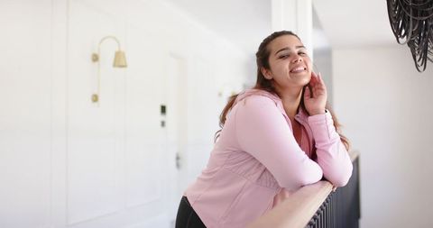 Smiling young woman leaning on banister in bright modern hallway wearing pink hoodie