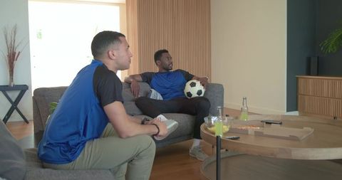 Diverse male friends watching soccer on sofa holding ball and snacking on pizza at home