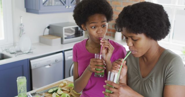 Mother and daughter sipping green smoothies in a modern kitchen, representing family bonding and healthy living. Ideal for illustrating concepts of nutrition, wellness, family time, healthy lifestyle, and homemade recipes. Could be used for blog posts, articles on family health, social media campaigns promoting nutrition, or advertisements for kitchen appliances or health foods.