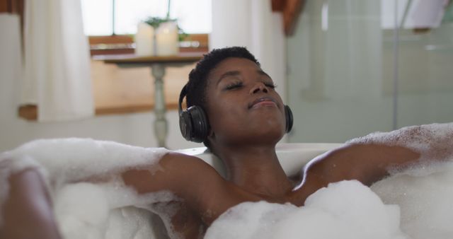 A young woman is relaxing in a bubble bath with closed eyes and wireless headphones, enjoying a moment of tranquility. Great for promoting self-care, relaxation routines, spas, wellness products, and leisure activities.