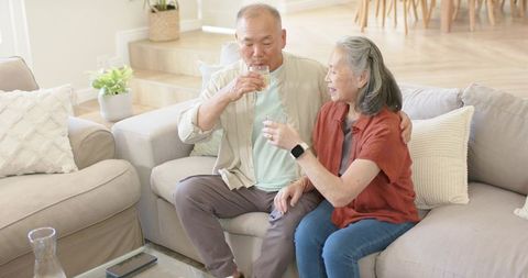 Asian senior couple relaxing on sofa sipping water in bright modern living room
