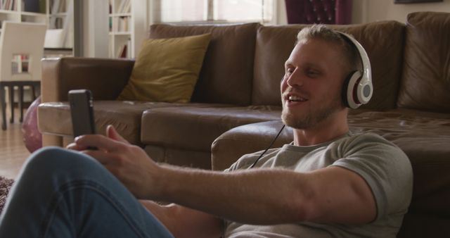 This image depicts a man sitting on the floor by a sofa, wearing headphones and using his smartphone for a video chat. He appears to be enjoying a leisurely moment at home. Ideal for illustrating home comfort, modern technology use, relaxation, work from home setups, remote communication, and personal time. Suitable for articles or advertising related to headphones, smartphones, home interior design, and communication technology.