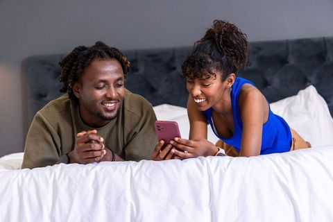 African american couple sharing smartphone and laughing on cozy bed, intimate bedroom moment