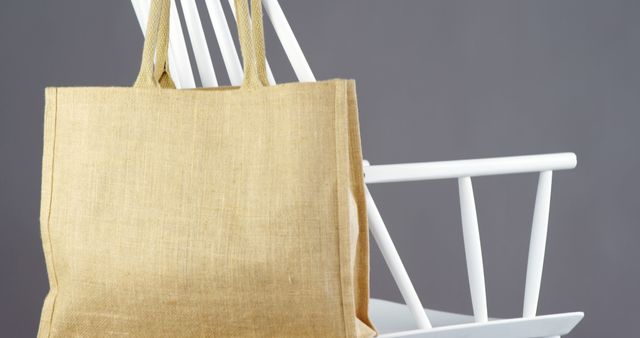 A beige tote bag hangs on the back of a white chair, with copy space. The simplicity of the scene suggests a focus on sustainable and eco-friendly fashion accessories.