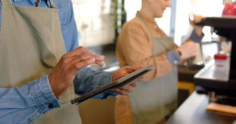 Baristas using tablet POS while steaming milk at cafe counter, teamwork in coffeehouse