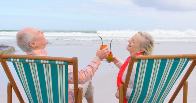 Senior couple smiling and enjoying drinks while sitting on striped beach chairs by the ocean. Great for retirement planning, senior lifestyle promotions, vacation brochures, or wellness advertisements focusing on leisure and happiness in later life.