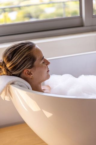 Woman relaxing in bubble bath by window, sunlit minimalist freestanding tub luxury retreat