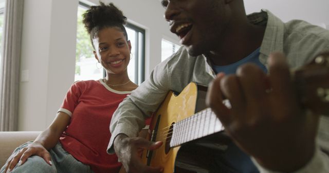 African American couple relaxing at home while one plays guitar and the other smiles and listens. Perfect for ads promoting music, relationships, home life, leisure activities, and modern living.
