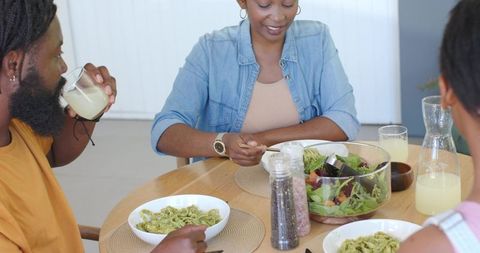 African american family sharing pesto pasta and garden salad around sunlit wooden table