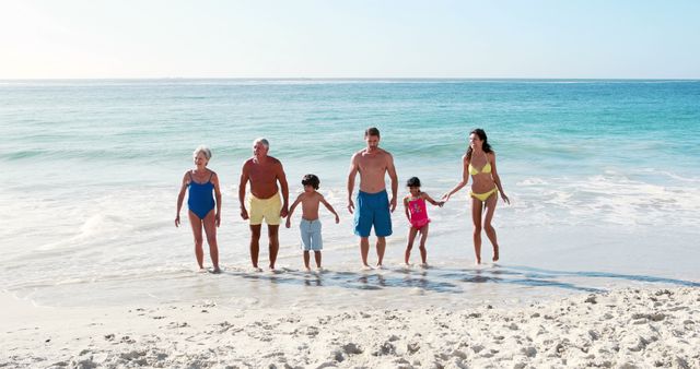 Multi-generational family forming a line holding hands walking on sandy beach near ocean. Ideal for use in travel brochures, advertisements promoting family vacations, articles on family bonding and summer activities, or wellness and lifestyle magazines.