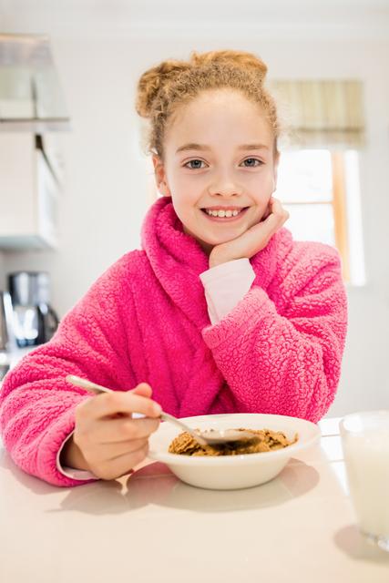Young girl in a bright pink bathrobe enjoying breakfast in a modern kitchen. She is smiling and looking at the camera while holding a spoon and eating from a bowl. This image is perfect for promoting healthy eating habits, family lifestyle, morning routines, and cozy home environments.